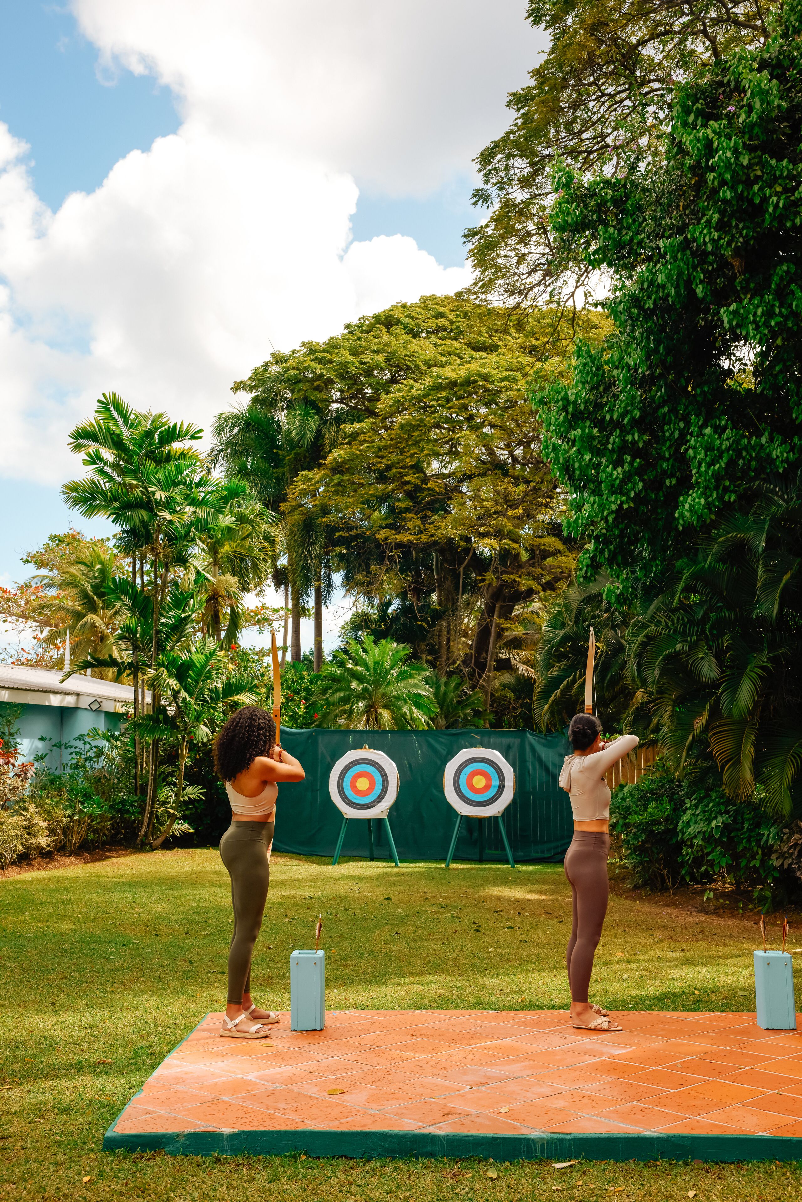 Two people practicing archery in an outdoor setting with targets and a green backdrop.