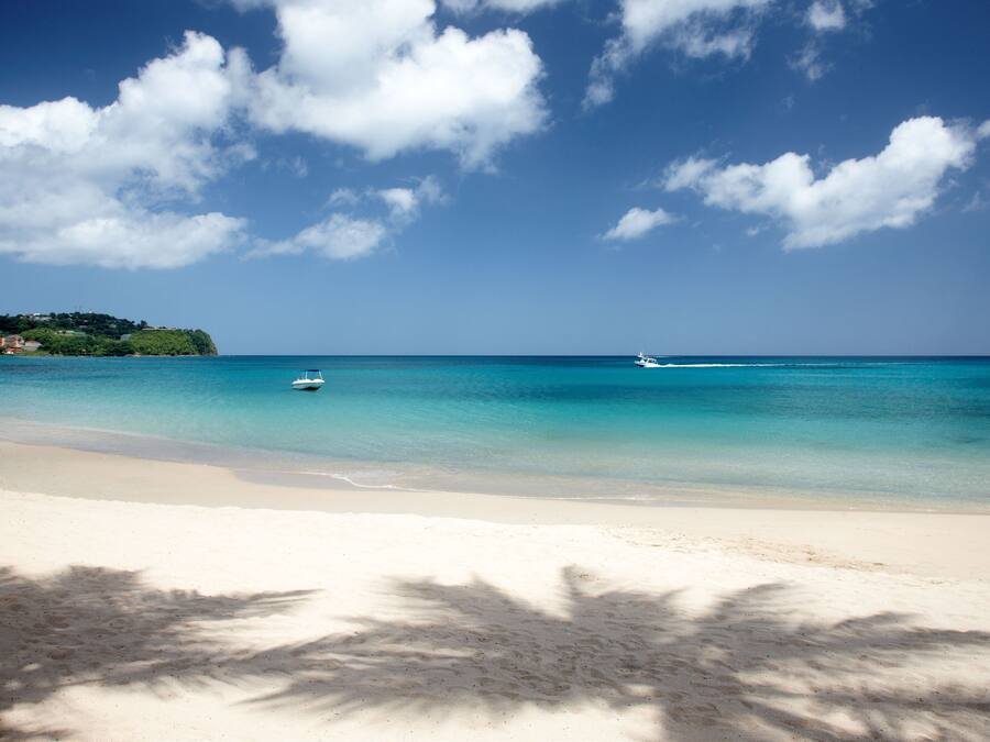 A beach with clear turquoise water, a boat in the distance, and a sandy shore with palm tree shadows.