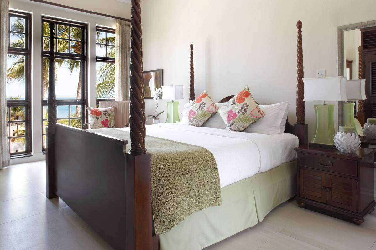 A four-poster bed with a green bed skirt, a nightstand with a lamp, and a large window with a view of palm trees.