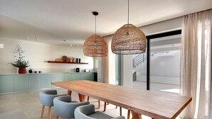 Un moderno comedor con una mesa de madera y cuatro sillas grises, dos lámparas colgantes de mimbre y una cocina al fondo.