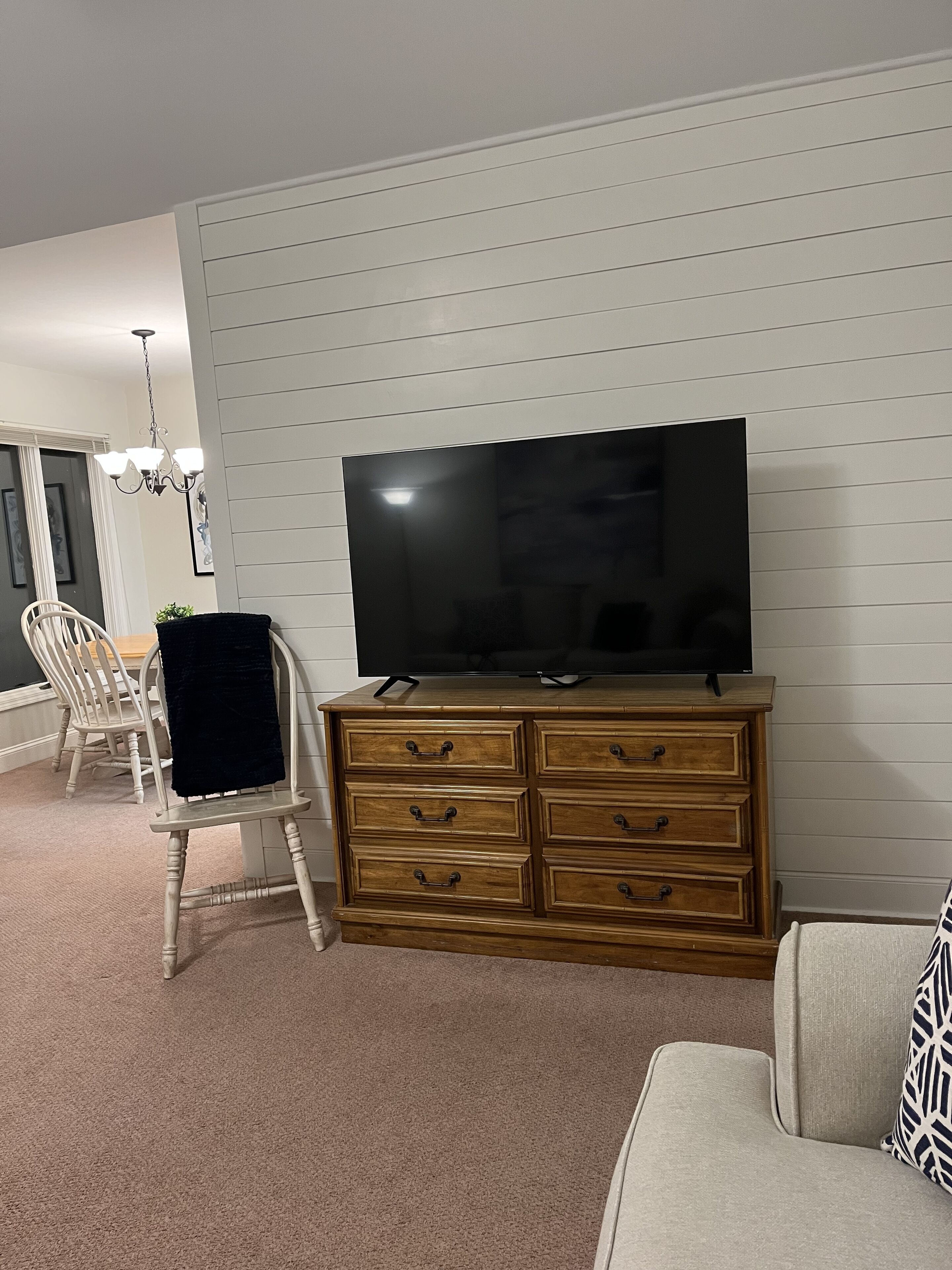 A living room with a wooden TV stand, a flat-screen TV, a white sofa, and a dining area with a chandelier.