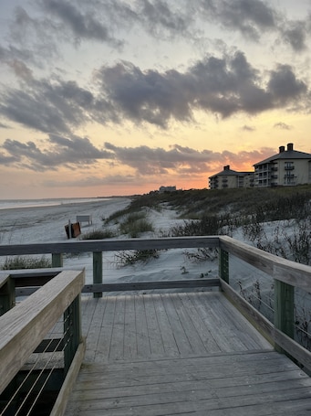 A wooden boardwalk leading to a beach with dunes and a building in the background.