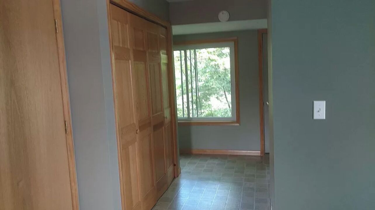 A hallway with a wooden door, a window with a view of trees, and a tiled floor.