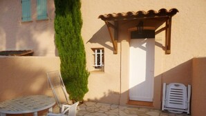 A patio with a stone floor, a white table with chairs, a tall green hedge, and a white door with a small window.