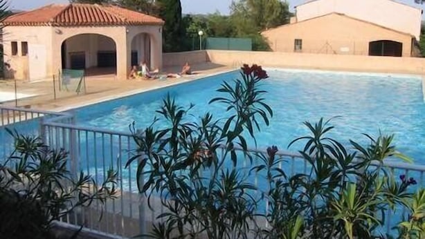 A swimming pool with a tiled deck, surrounded by palm plants and a building with a tiled roof.
