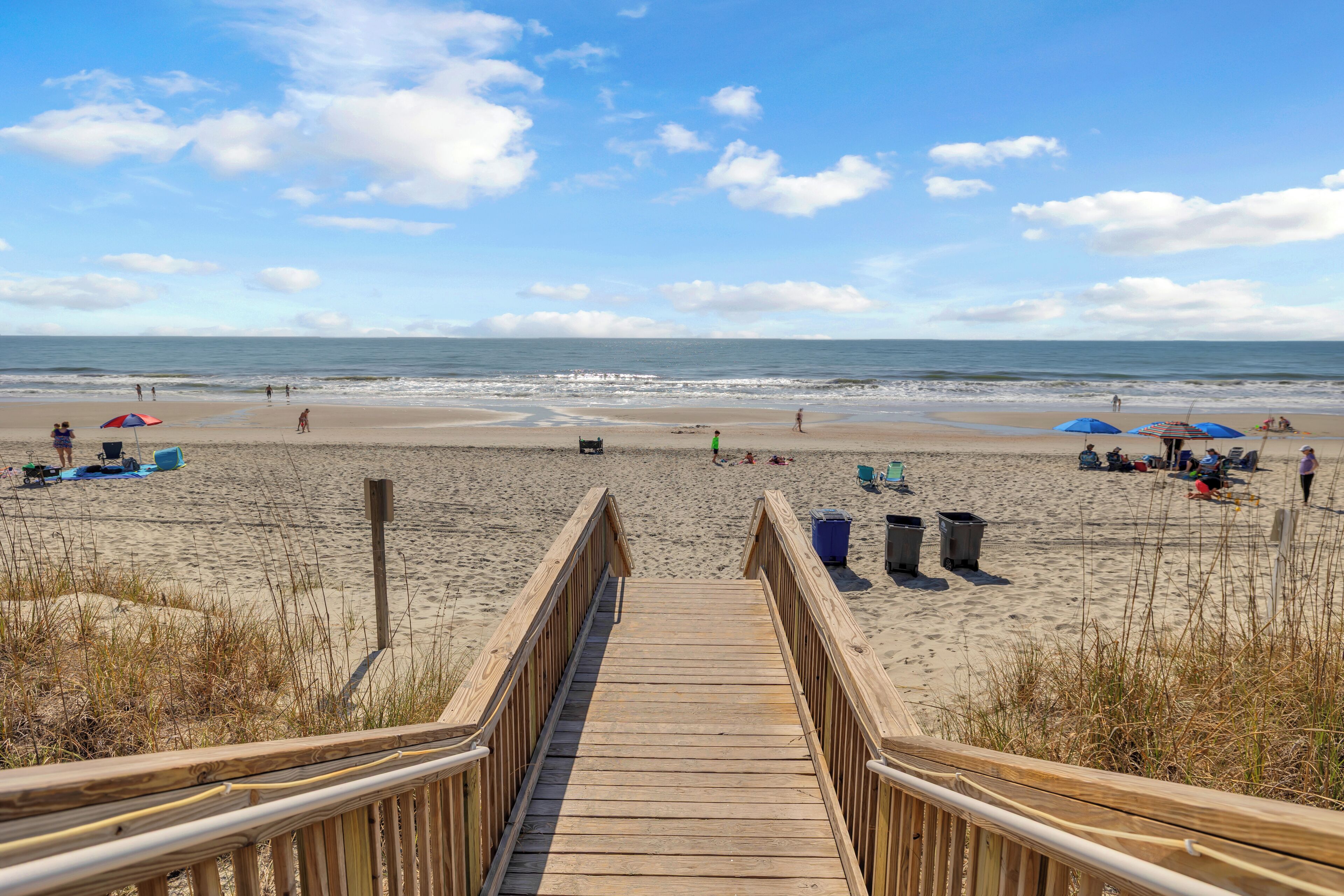 A wooden boardwalk leading to a beach with umbrellas and people.