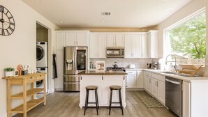 A modern kitchen with white cabinets, stainless steel appliances, a wooden bar stool, and a large window. - Eastside Escape *backyard games, Walk to Bars, Brews & Good Eats* (Austin)
