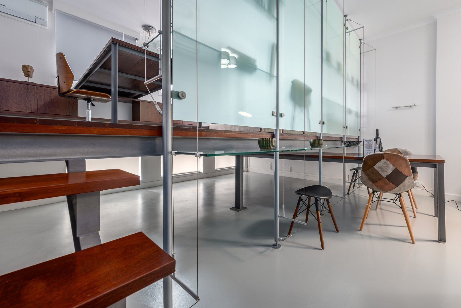 Een modern interieur met een glazen wand, houten banken, een metalen tafel en een stoel met een geometrisch kussen.