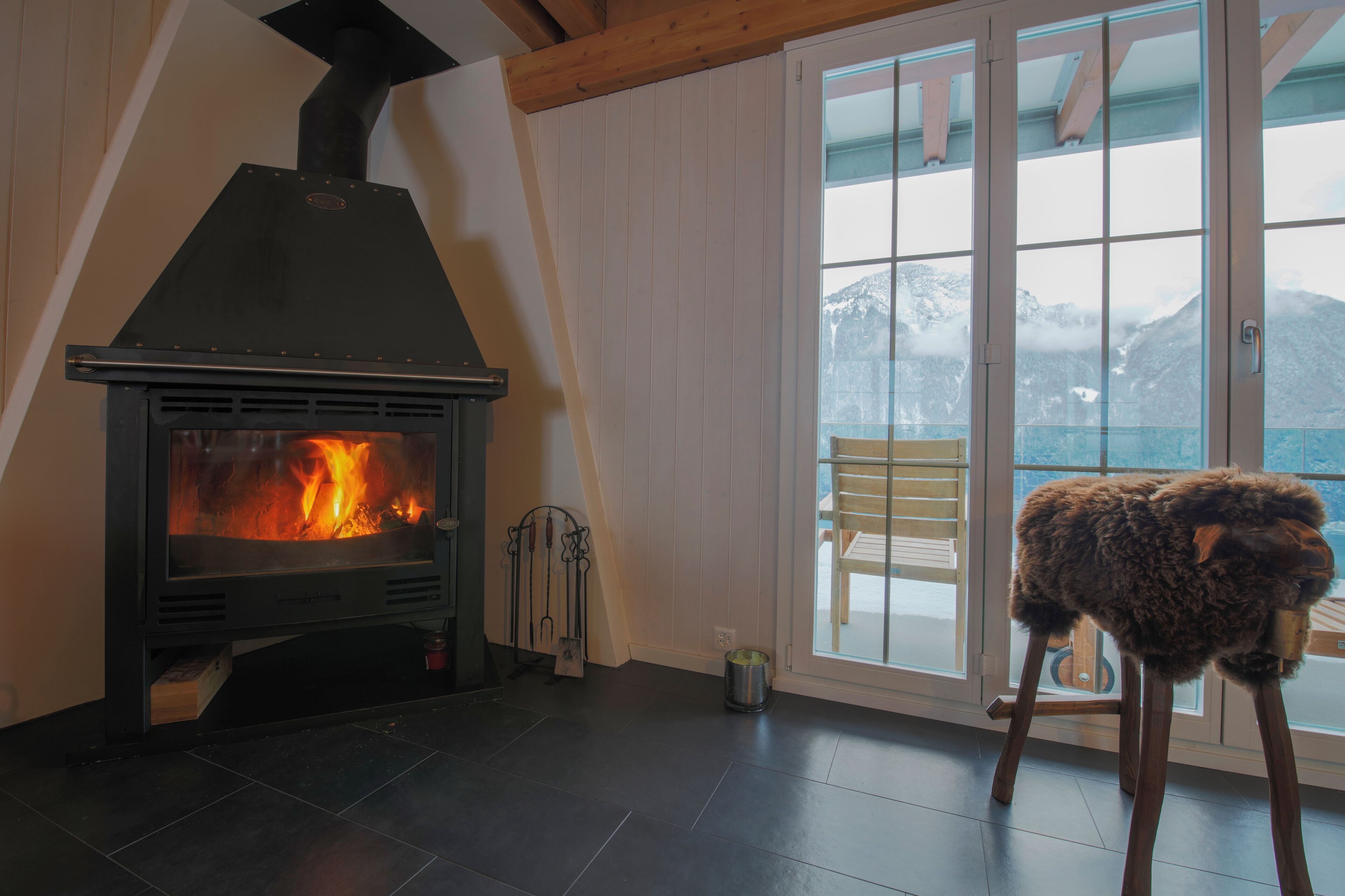 Ein Zimmer mit Kaminofen, einem Holzstuhl und Bergblick durch eine Glastür.