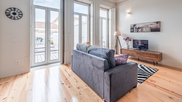 Ein modernes Wohnzimmer mit einem grauen Sofa, einem Holzschrank und einem großen Fenster mit Blick auf Gebäude.
