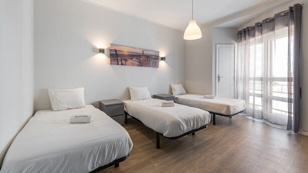 Habitación de hotel con tres camas individuales, un cuadro en la pared, una lámpara colgante y un ventanal con cortinas.