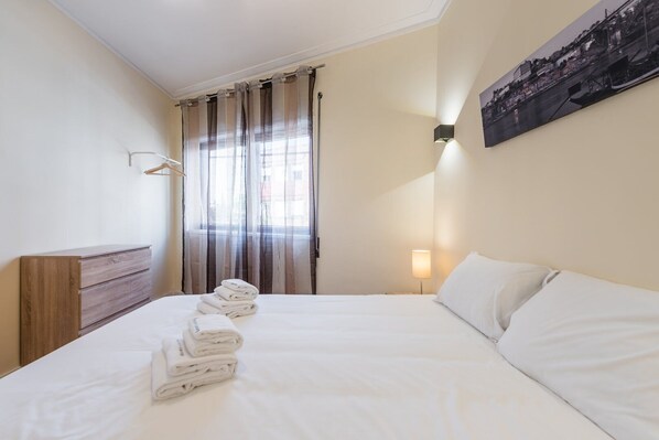 A hotel room with a neatly made bed, a wooden dresser, a window with curtains, and a wall-mounted lamp.