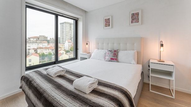 A modern bedroom with a large window, a bed with white linens, a striped rug, and two bedside tables with lamps. - GuestReady - Cosy spot near Porto city centre (Porto)