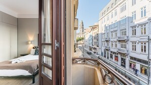 A hotel room with a bed, a nightstand, and a window overlooking a street with buildings and a clock tower. - GuestReady - Rivoli Studio (Porto)