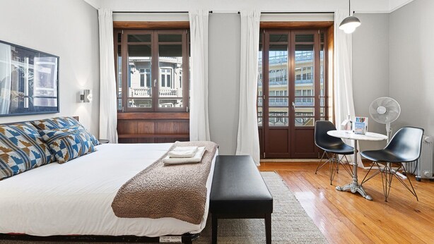 A modern hotel room with a large bed, a small table with two chairs, a standing fan, and a view of the city through large windows. - GuestReady - Rivoli Studio (Porto)