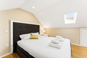 A neatly made bed with white linens and a single yellow pillow, a bedside lamp, and folded towels on the floor.