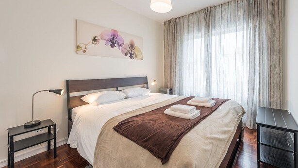 A neatly made bed with white linens and a brown throw blanket, a bedside table with a lamp, and a large window with sheer curtains.