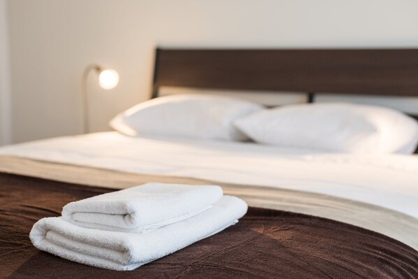 A neatly made bed with white linens and pillows, a folded white towel on the bed, and a bedside lamp.