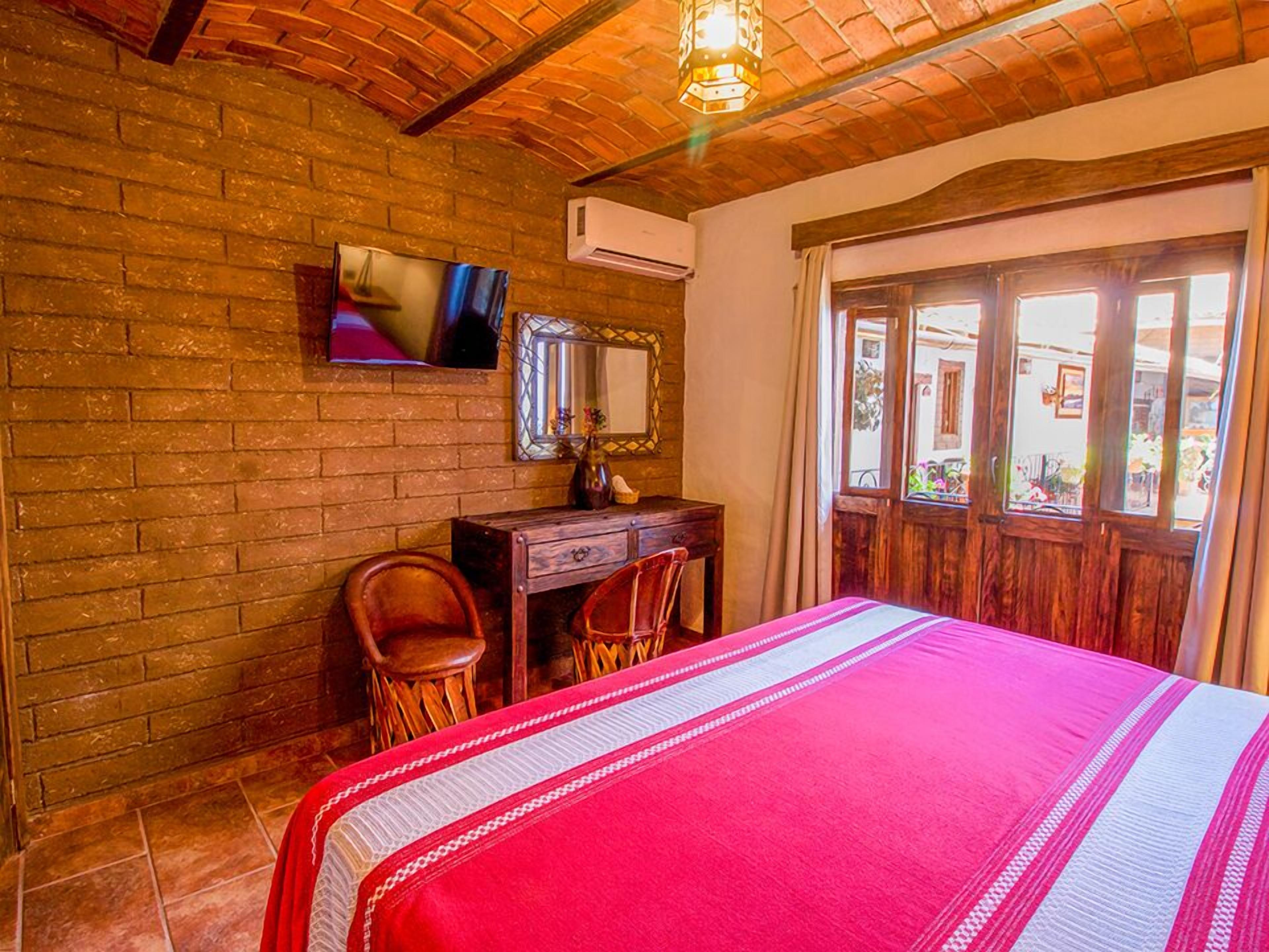 A room with a brick wall, a wooden desk with two chairs, a bed with a red and white striped bedspread, a TV mounted on the wall, and an air conditioning unit.