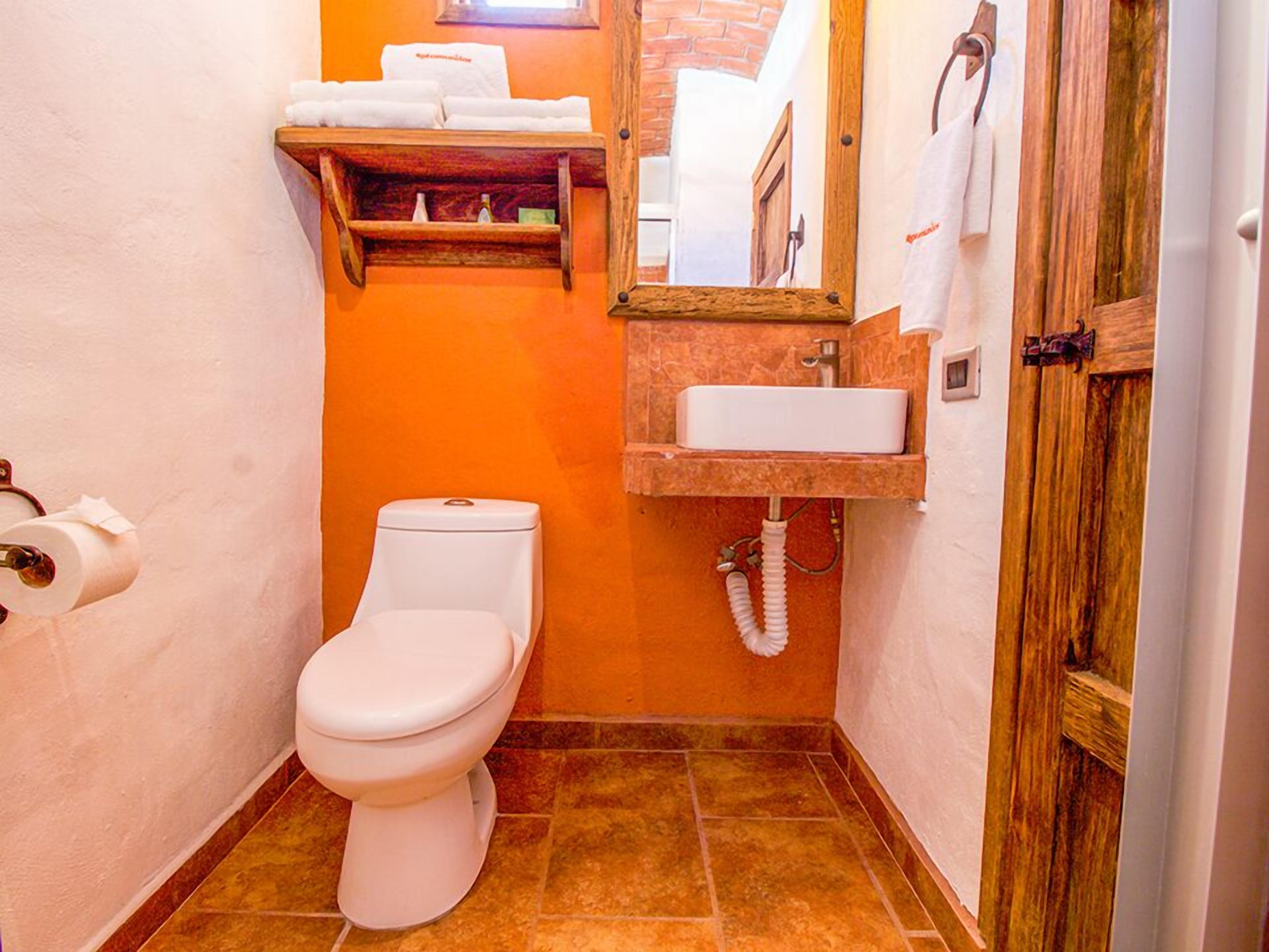 A compact bathroom with a white toilet, a wooden shelf with towels, a sink with a mirror, and a door with a towel hanging.
