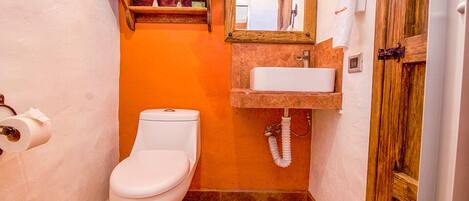 A compact bathroom with a white toilet, a wooden shelf with towels, a sink with a mirror, and a door with a towel hanging.
