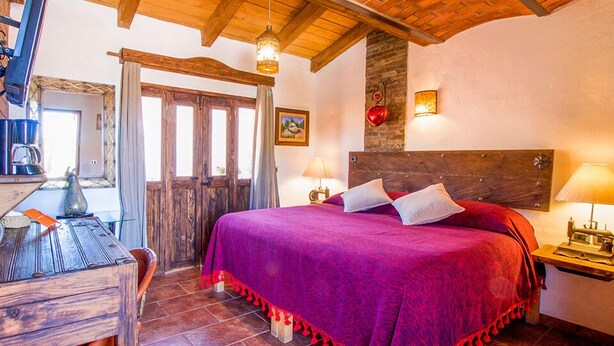 A cozy bedroom with a wooden ceiling, a large bed with a purple bedspread, a wooden desk with a lamp, and a stone wall with a heart decoration. -  Hotel Mi Pueblito Tapalpa (Tapalpa)