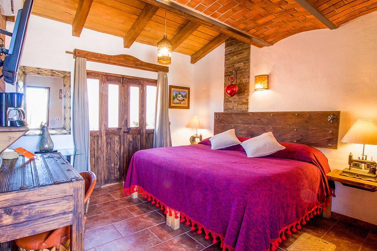 Een gezellige slaapkamer met een houten plafond, een groot bed met een paars dekbed, een houten bureau met een lamp en een stenen muur met een hartdecoratie.