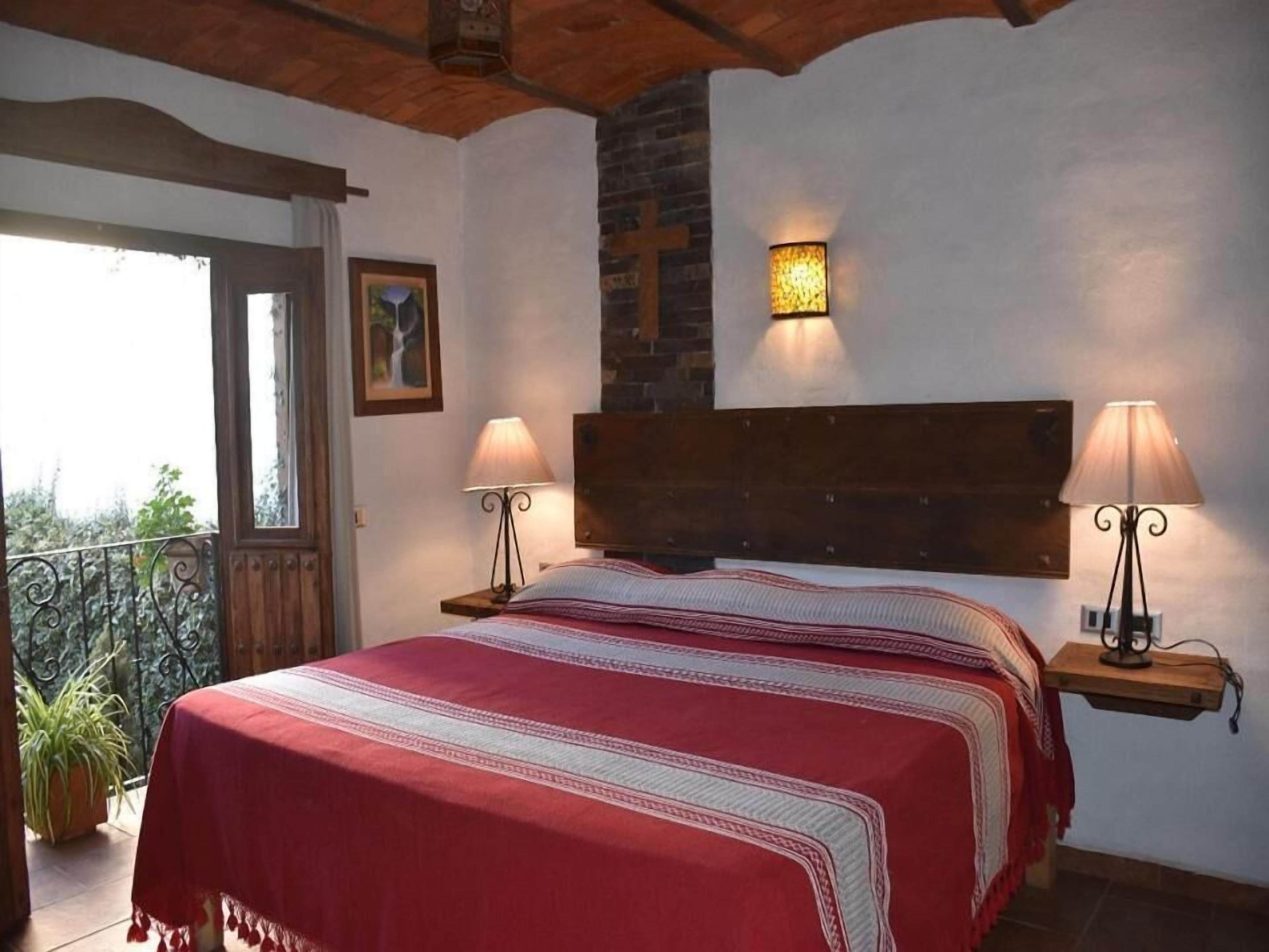 Ein Schlafzimmer mit einem großen Bett, zwei Nachttischen mit Lampen, einem Holz-Kopfende, einer Ziegelwand und einem Balkon mit Geländer und Topfpflanze.