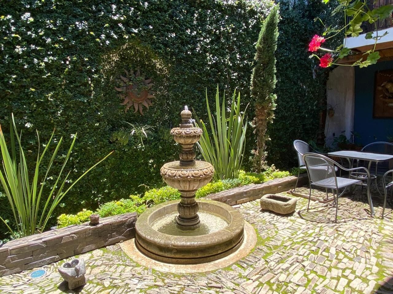 A stone fountain in a garden with a stone wall and a table with chairs.