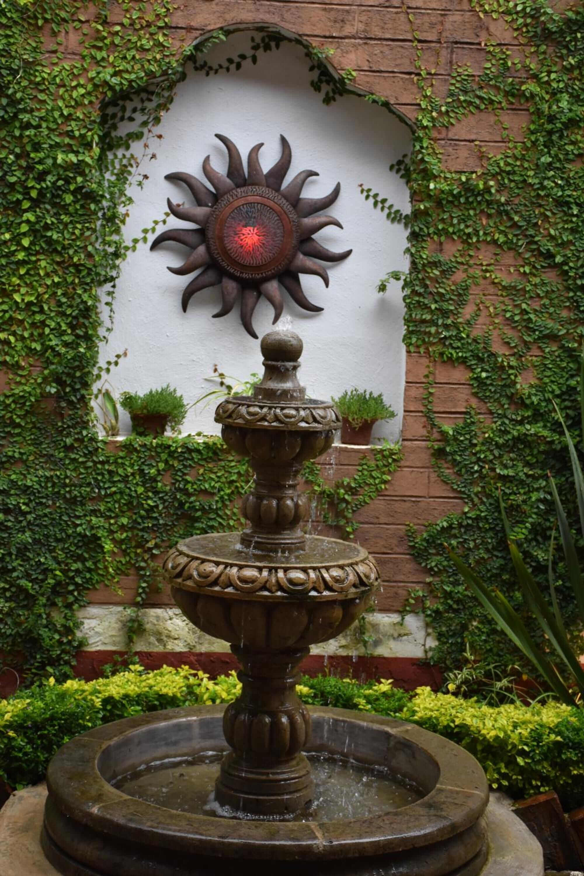 A fountain with a decorative sun in the background, surrounded by greenery and potted plants.