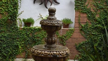 A fountain with a decorative sun in the background, surrounded by greenery and potted plants.