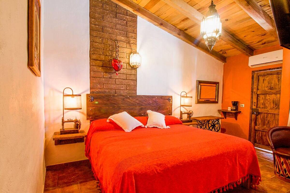 Een knus ingerichte kamer met een groot bed, een houten hoofdbord en een accentmuur van baksteen. Er zijn twee nachtkastjes met lampen en een klein tafeltje met een kaars.