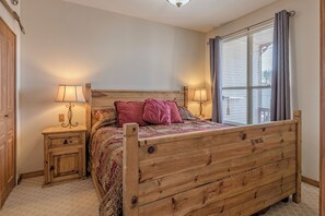 A bedroom with a wooden bed, two bedside tables with lamps, a window with curtains, and a door.