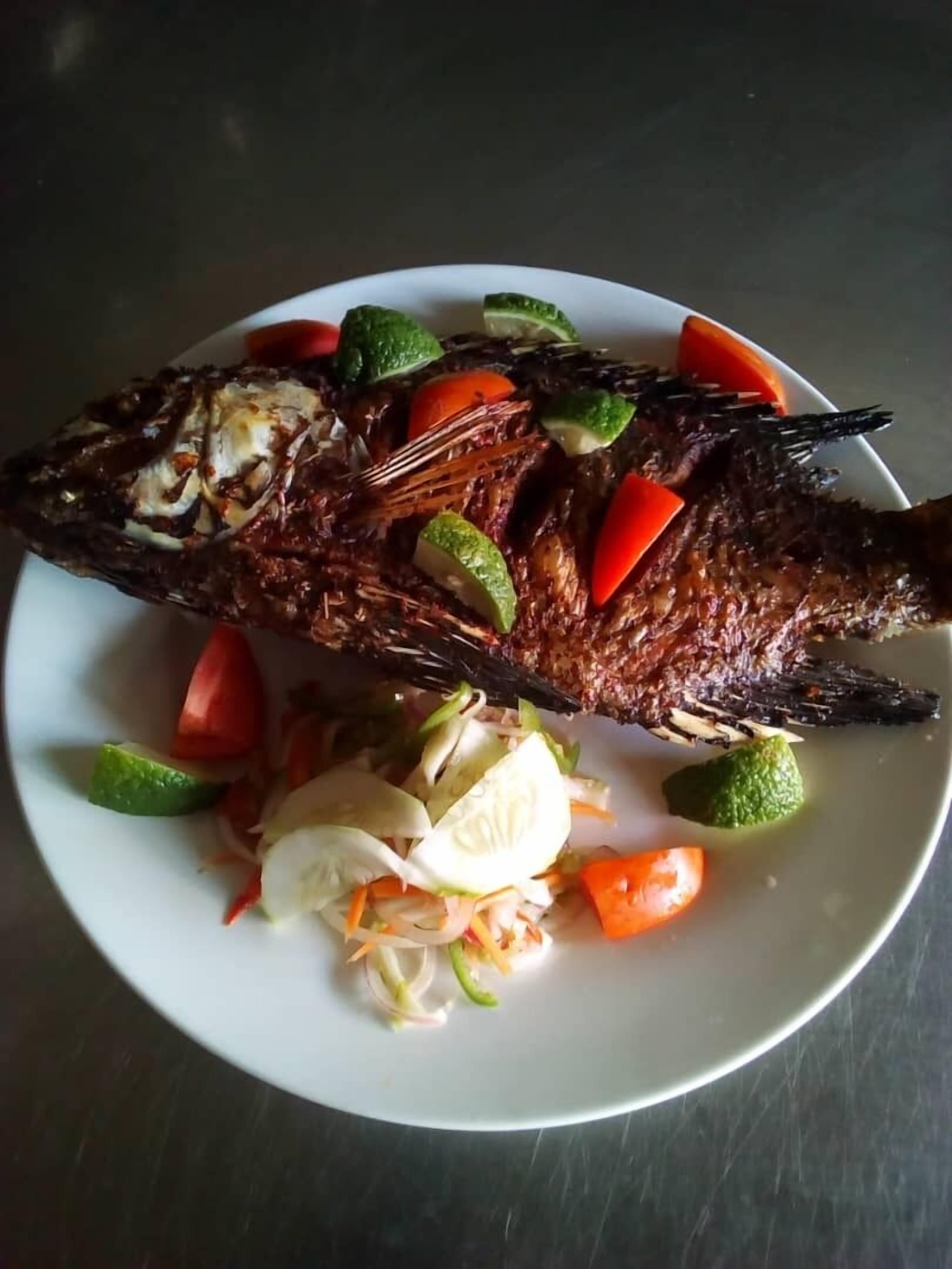 A whole roasted fish on a plate with sliced vegetables and lime wedges.