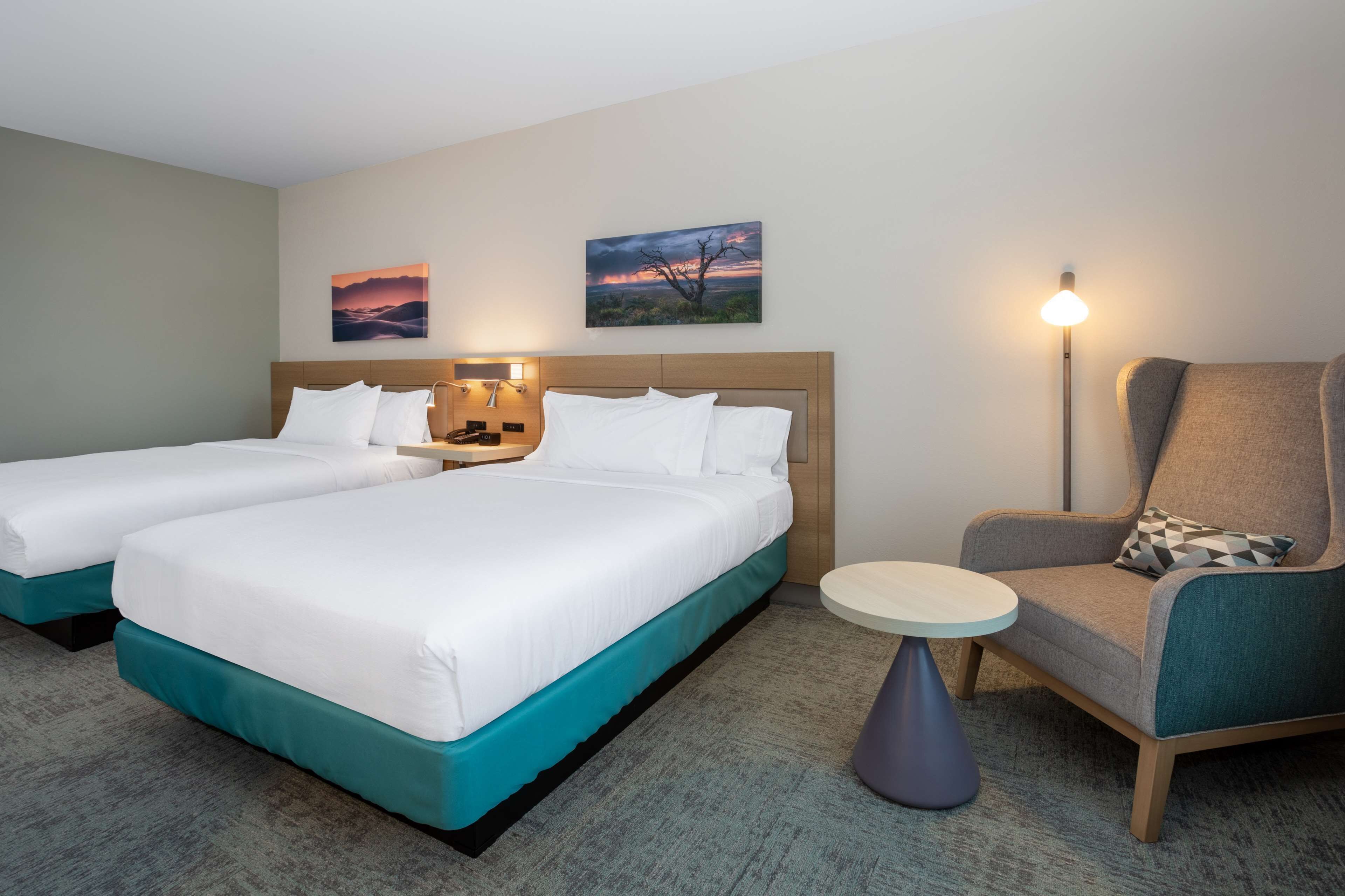 Habitación de hotel con dos camas, una silla, una mesa y dos cuadros colgados en la pared.