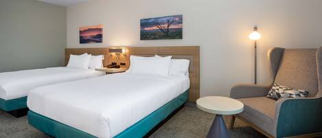A hotel room with two beds, a chair, a table, and two wall-mounted paintings.