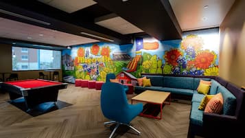 Una mesa de billar, un sillón azul, un sofá con cojines de colores y un mural con flores y un edificio.