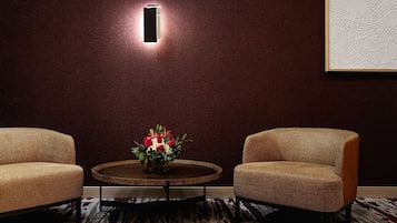 A room with two wicker chairs, a round wooden table with a vase of flowers, a wall-mounted light, and a textured wall.