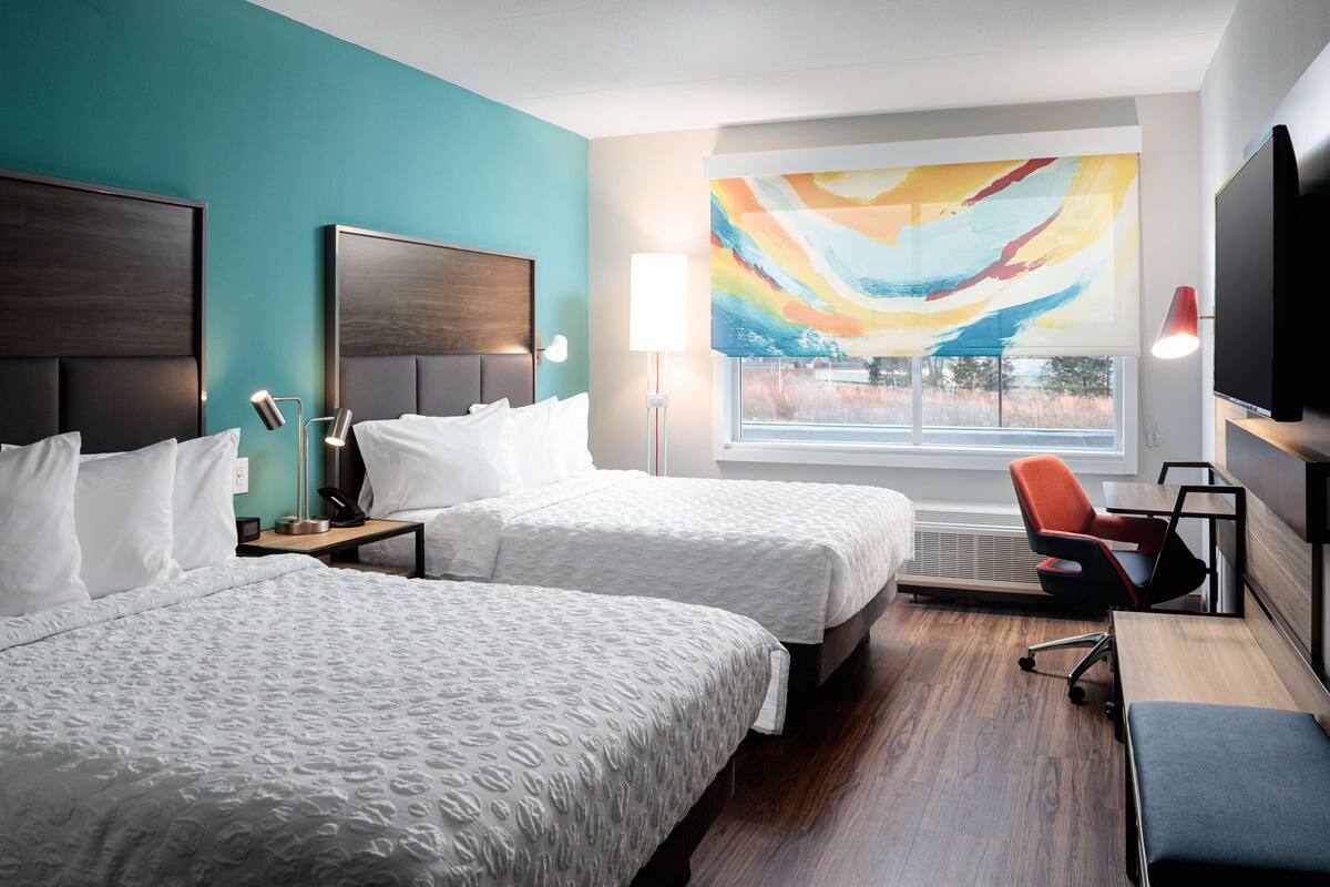 A hotel room with two beds, a desk, a chair, a large window with a view, and a colorful abstract painting.