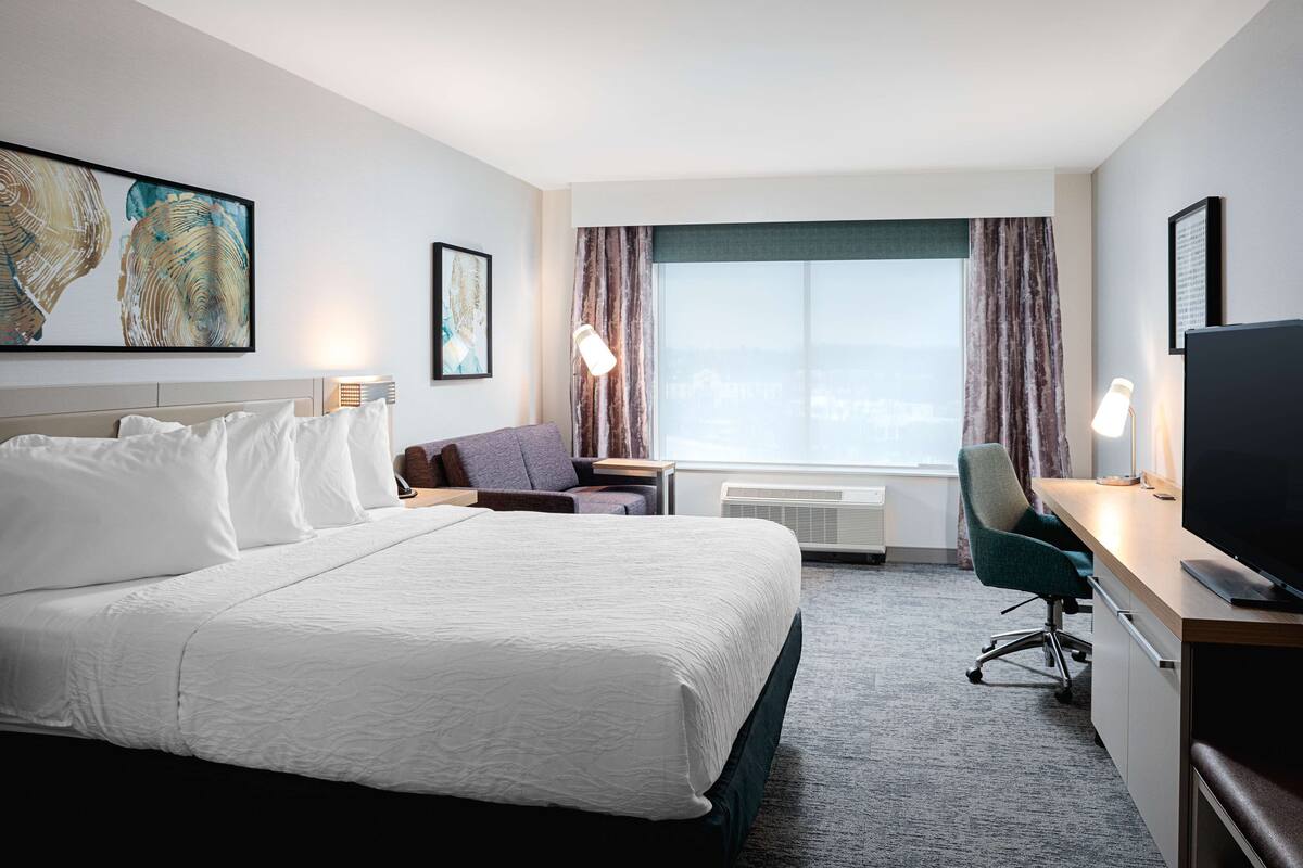 A hotel room with a large bed, a desk with a computer, a chair, and a TV. There is a window with curtains and a view of the city.