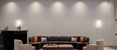 A modern hotel lobby with a sectional sofa, two armchairs, a small table, and a bar area with bottles and glasses.