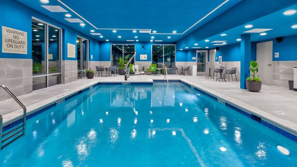 Una piscina con piastrelle blu, circondata da sedie e tavoli, e un cartello che avvisa dell'assenza di bagnino.