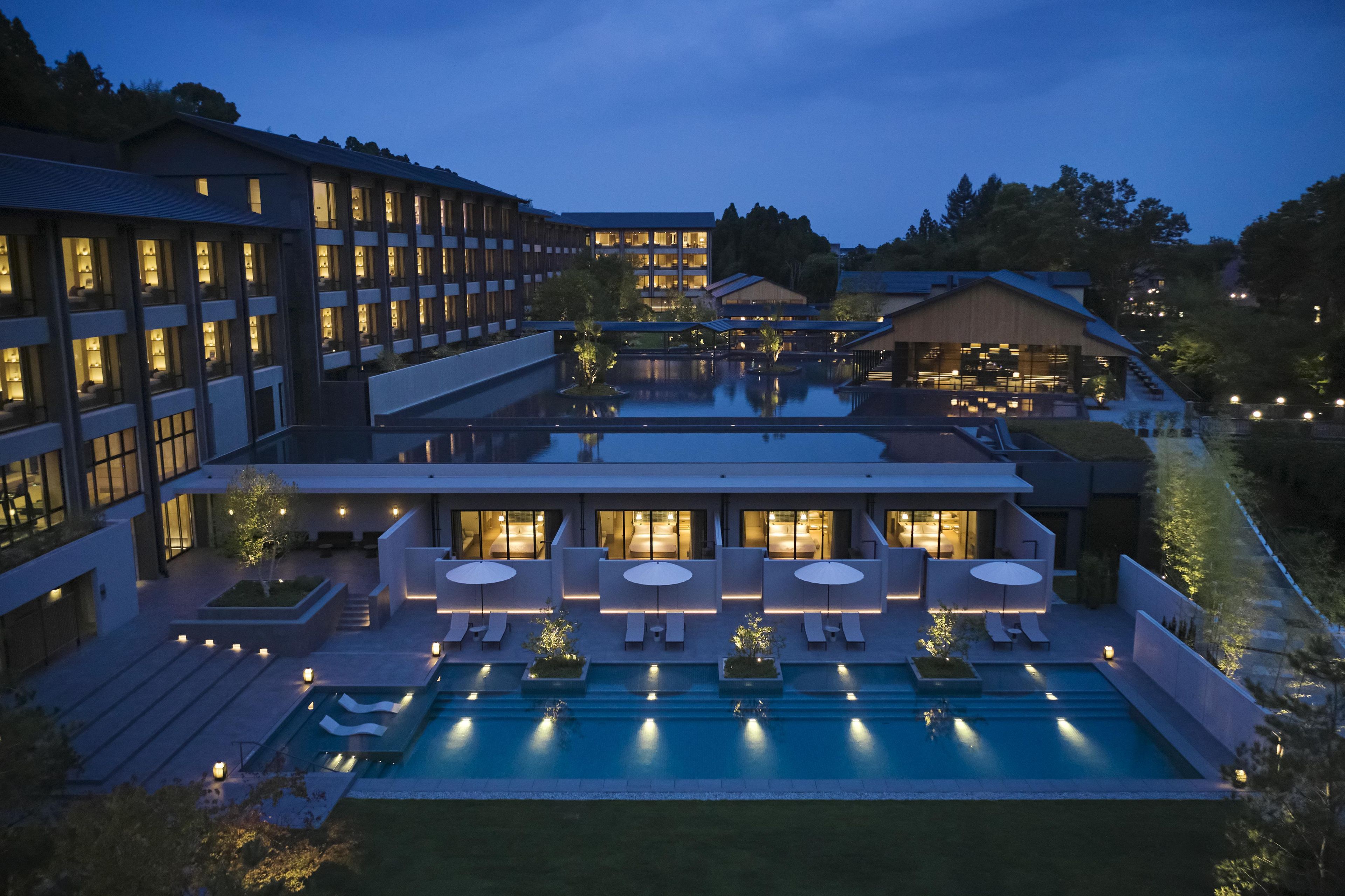A modern hotel with multiple swimming pools, outdoor seating areas, and a well-lit facade.