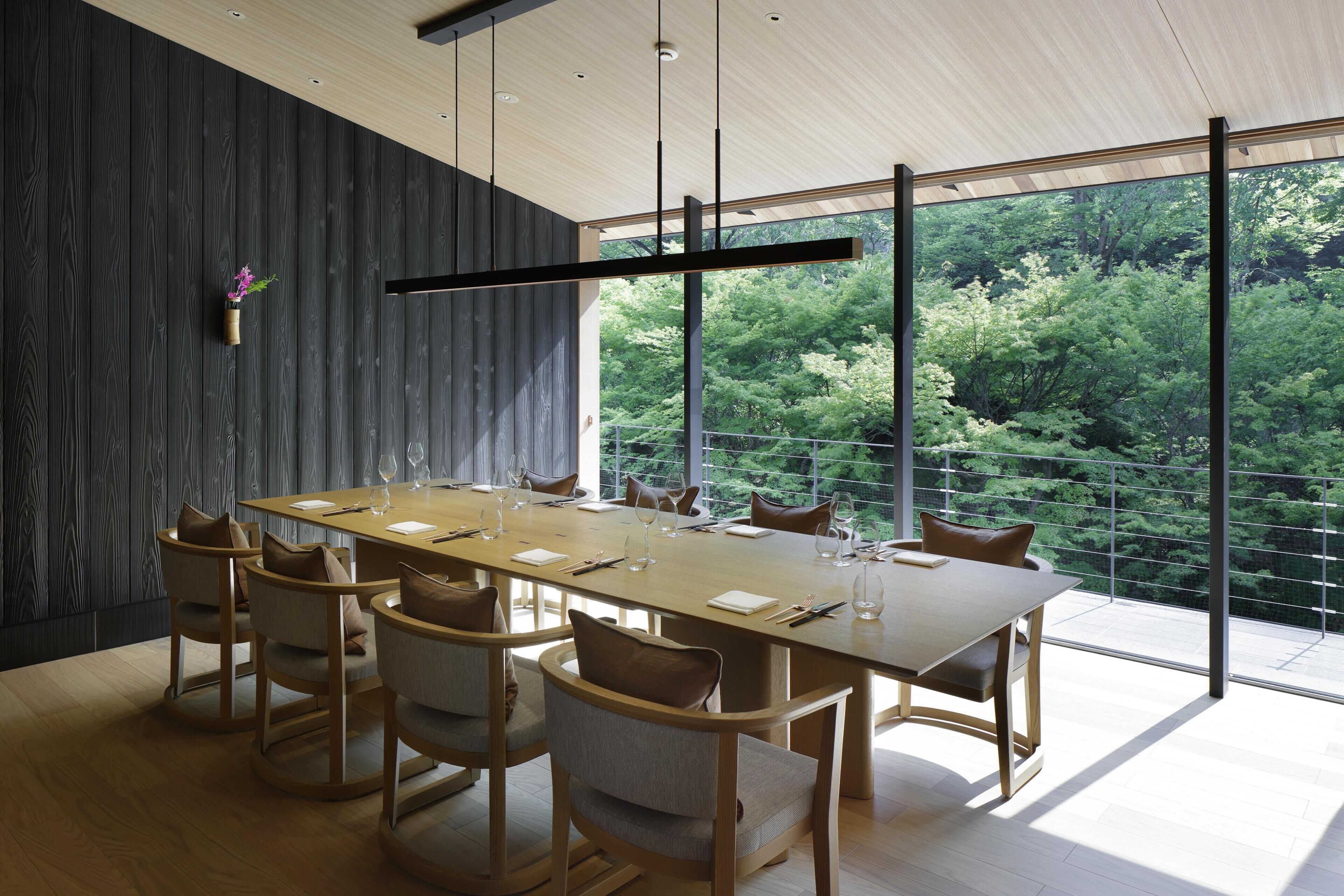 A modern dining room with a long table, chairs, and a view of a forest through large windows.