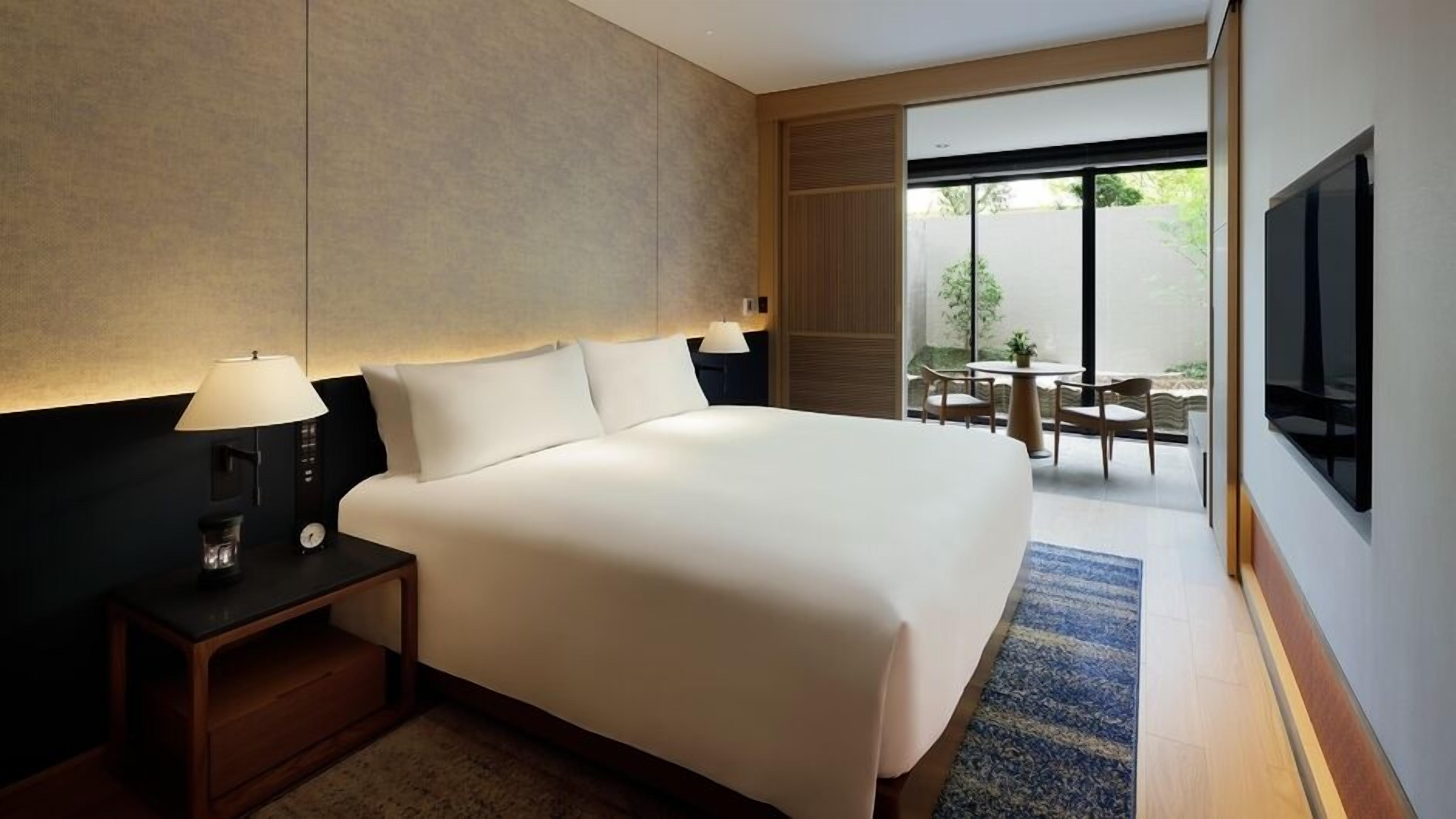 Een hotelkamer met een groot bed, een nachtkastje met een lamp, een televisie aan de muur en uitzicht op het buitengebied.