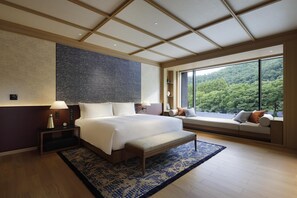 A modern hotel room with a large bed, a bench, a view of the forest, and a large window.