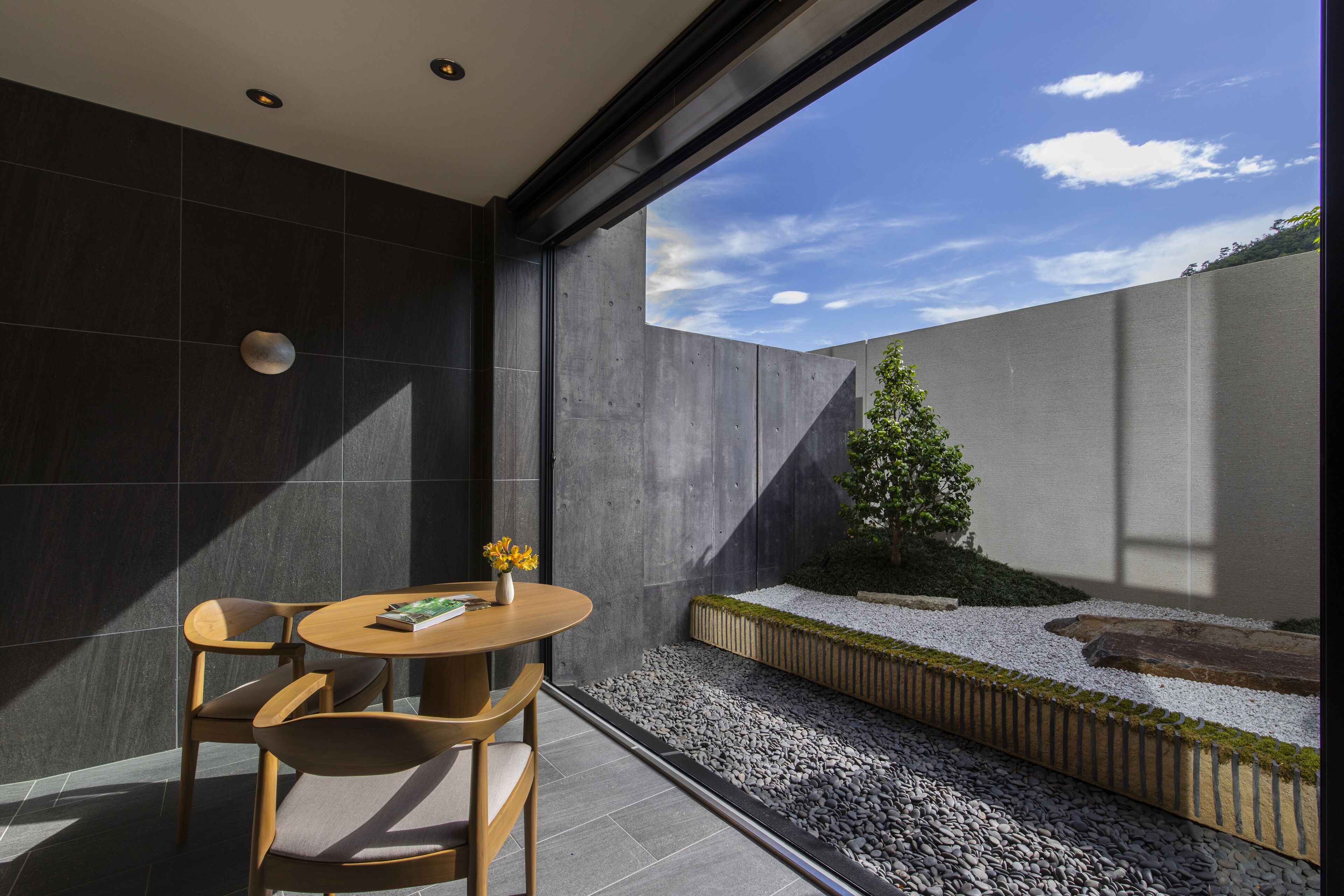 A modern interior with a dining area featuring a round table and two chairs, a small potted plant, and a view of a garden with a tree and gravel path.