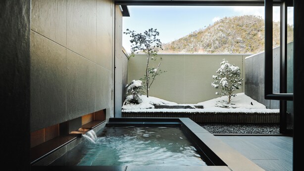 A modern spa area with a water feature, snow-covered plants, and a view of a hilly landscape.
