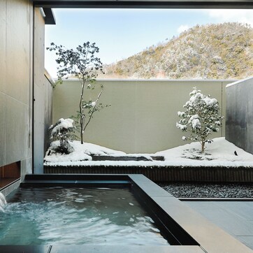 A modern spa area with a water feature, snow-covered plants, and a view of a hilly landscape.