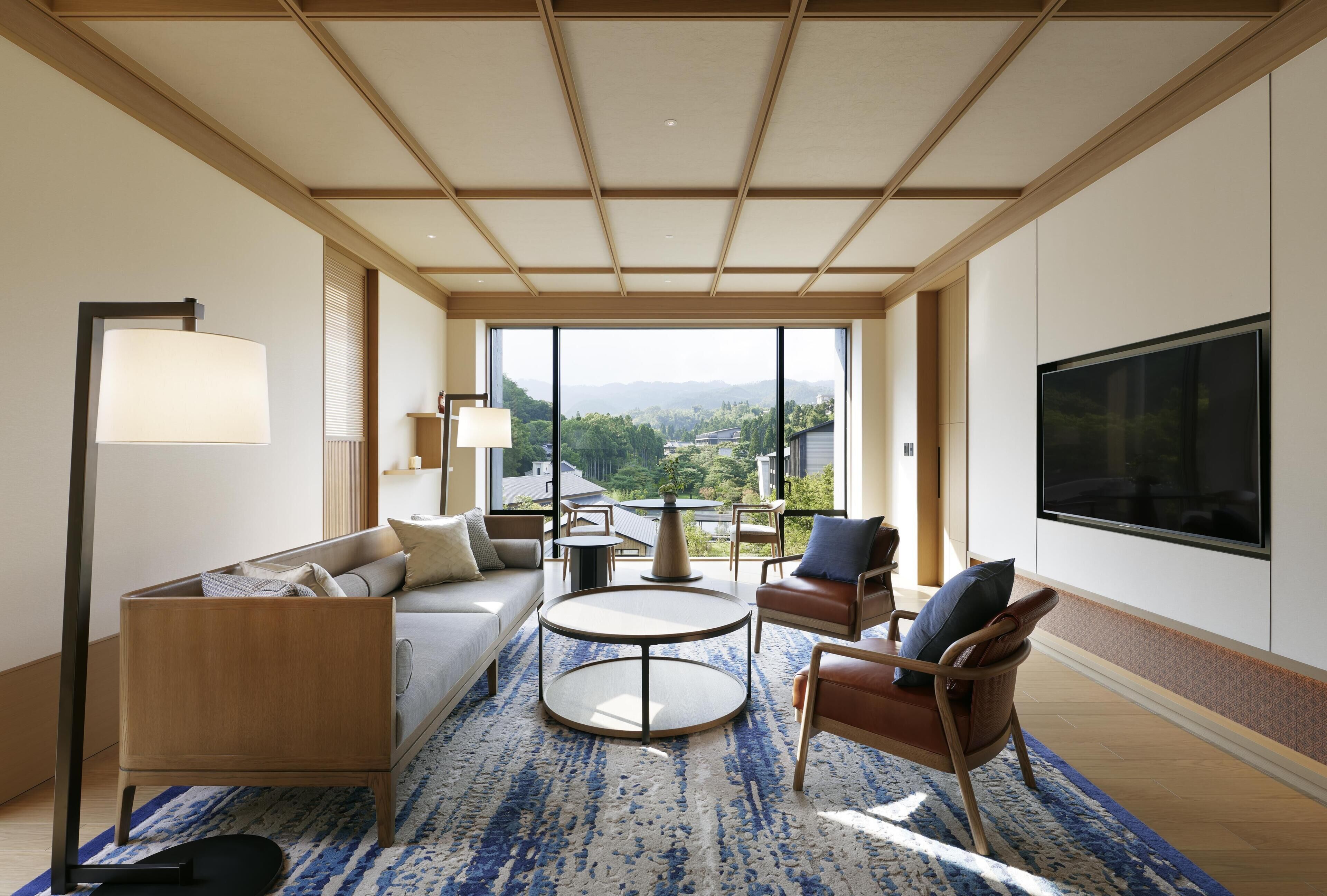 A modern hotel room with a sofa, chairs, a coffee table, a TV, and large windows offering a view of the outdoors.